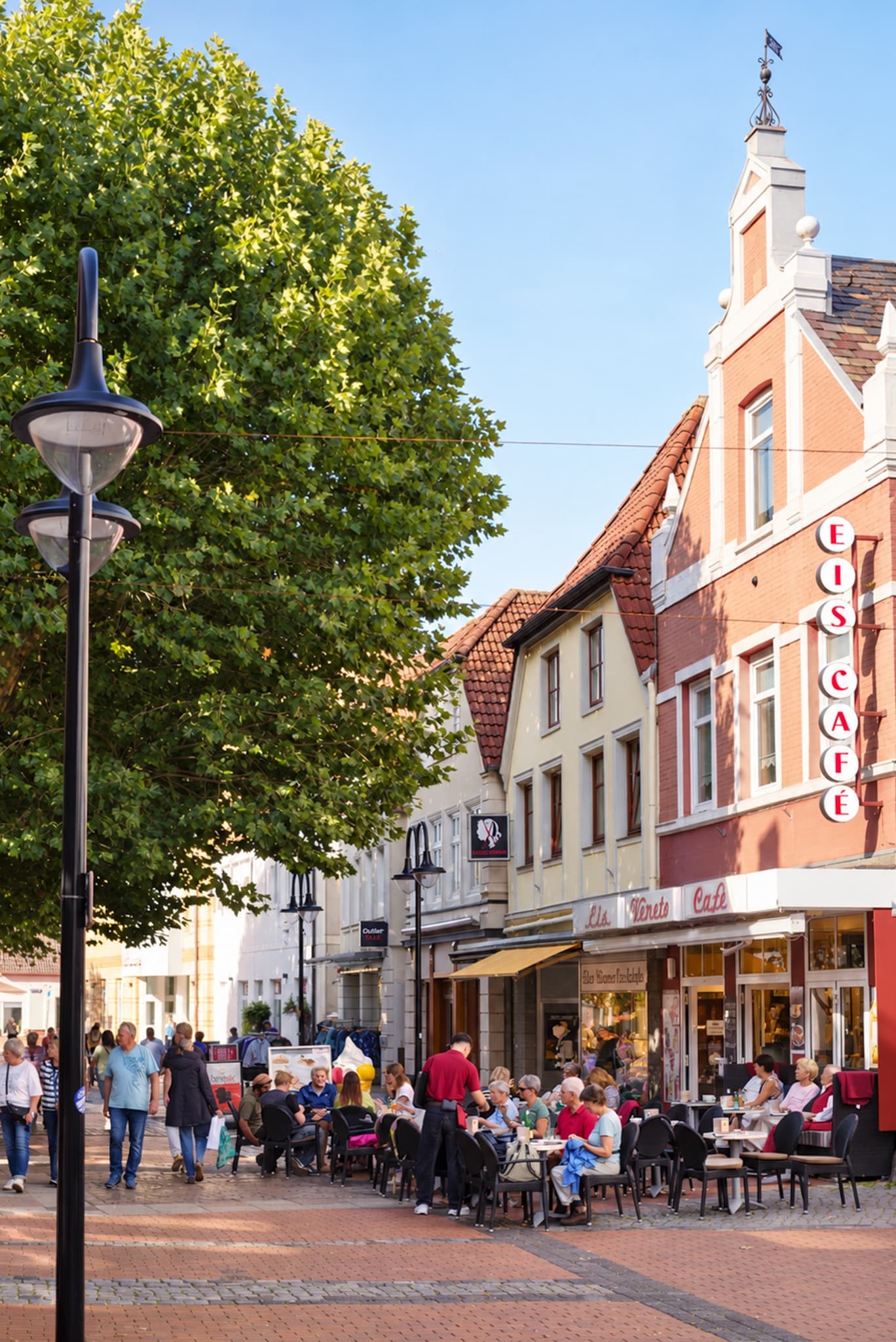 Die rote Giebelfassade in der Einkaufsmeile mit senkrechtem Schriftzug „Eis Café“ und dem Schild „Eis Veneto Café“, davor eine Laterne und ein Laubbaum
