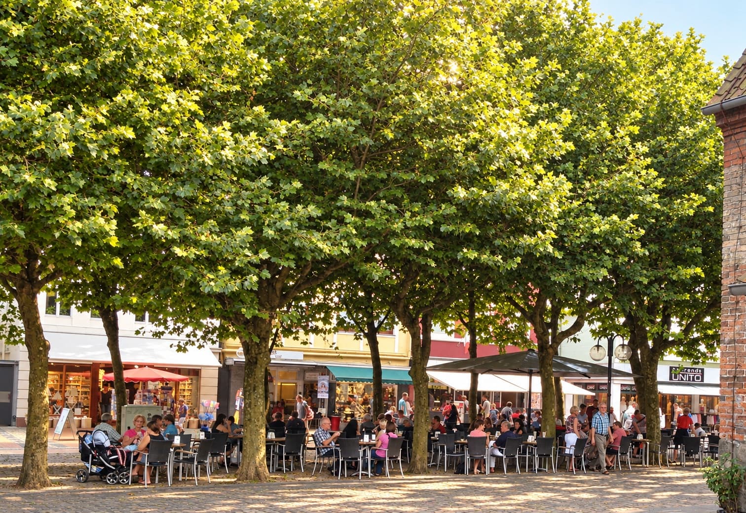 Weiter Blick über die Terrasse in der Einkaufsmeile unter großen Platanen, viele Gäste an den Tischen