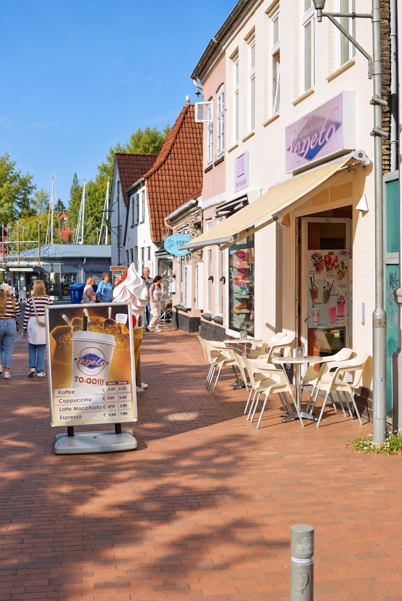 Eiscafé Veneto am Hafen — helle Fassade mit Veneto-Schild und Markise, davor Aufsteller und weiße Stühle, im Hintergrund Bootsmasten