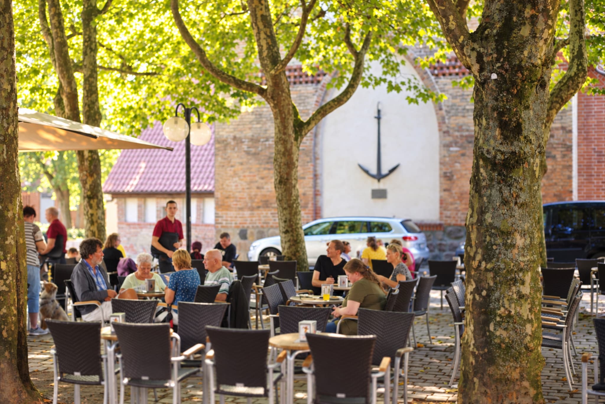 Außenterrasse in der Einkaufsmeile unter Platanen, im Hintergrund eine weiße Giebelwand mit Anker