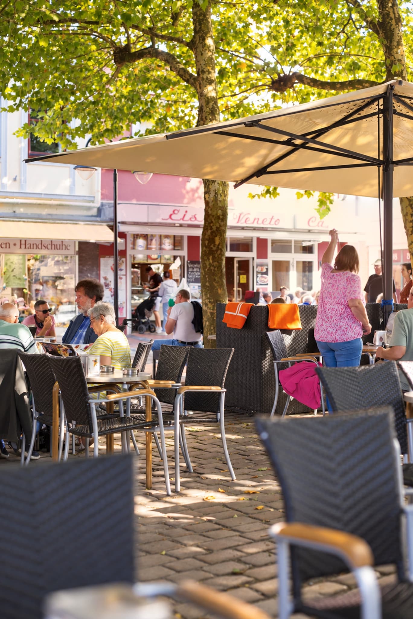 Gäste unter einem großen Sonnenschirm auf der Terrasse in der Einkaufsmeile, dahinter die rote Veneto-Fassade