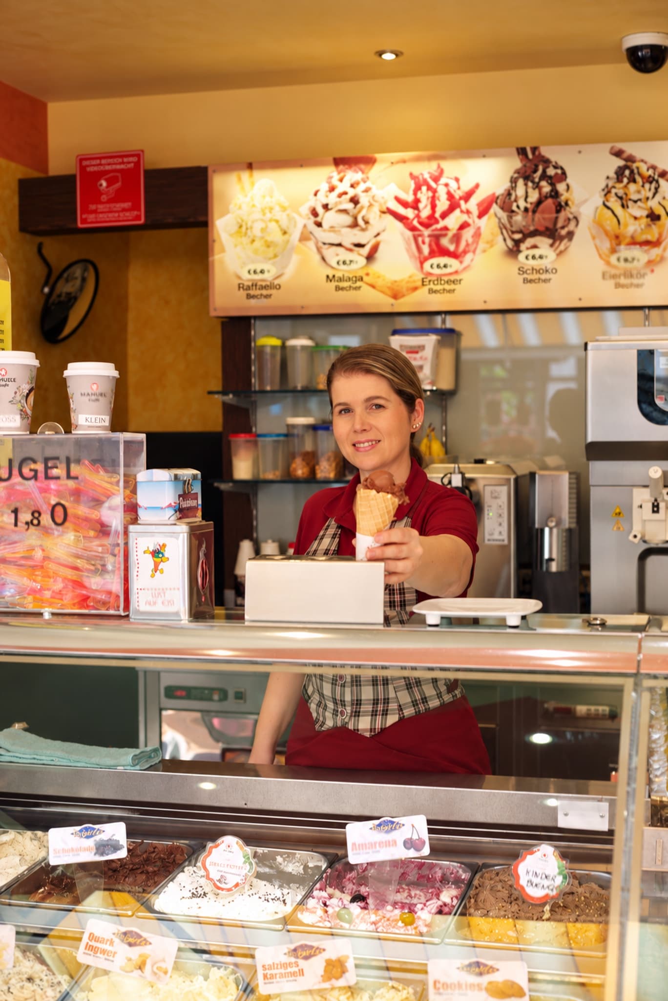 Am Hafen reicht eine Mitarbeiterin im roten Poloshirt lächelnd eine Eiswaffel über die gefüllte Gelato-Vitrine